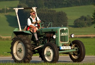 Traditionally dressed farmer in lederhosen and Tyrolean hat on a tractor, Dietramszell, Bavaria,