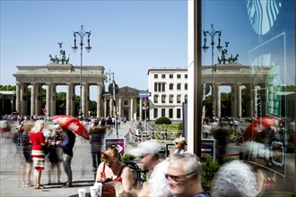 The Brandenburg Gate is reflected in a pane of glass. A long exposure shows tourists at Cafe am