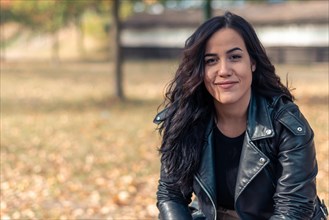 Content latina woman in a leather jacket with a relaxed posture in an autumnal park setting,