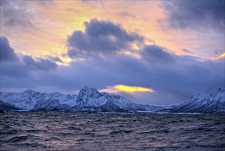 Sunset over a snowy mountain range by the ocean with a dramatic sky, Lofoten
