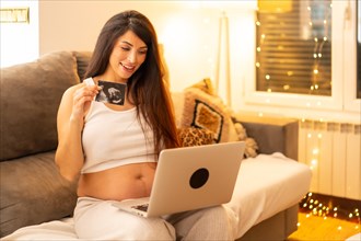 Happy pregnant woman showing ultrasound during online video call sitting on the sofa at home at