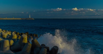 Evening view of a calm seashore with spray over tetrapods, lighthouse in the background, in South