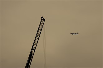 Aircraft in flight with an industrial crane in the foreground at sunset, London, England, United