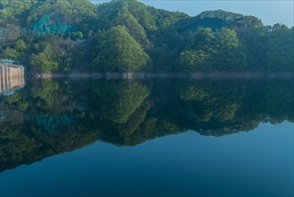 Calm reservoir with forested hills and morning mist, creating a mirror-like reflection, in South