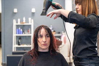 Frontal photo of a serious and patient woman sitting on hair salon while hairdresser drying her