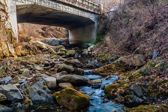 Leafless trees and rocks surround a stream flowing under a bridge in an autumn setting, in South