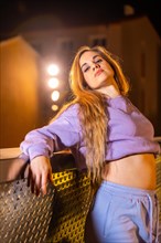 Vertical photo of a blonde beauty trap dancer posing leaning on a urban rail at night