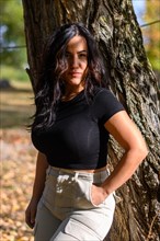 Confident hispanic sexy woman in black top and khaki pants standing by a tree in autumn, blurred