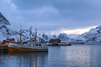 Docked fishing boats in a serene harbor with snow-covered mountains under the evening light,