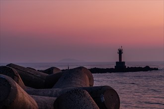 Dusk light casts an orange glow over a sea with a lighthouse and breakwater blocks, in South Korea