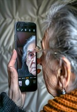 An elderly woman holds her smartphone in her left hand and looks at her reflection on the display,
