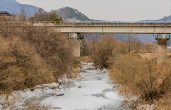 An overcast winter scene showing a bridge over a partially frozen creek with leafless trees, in