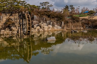 Old water wheel reflected in a pond surrounded by rocks and trees under a cloudy sky, in South