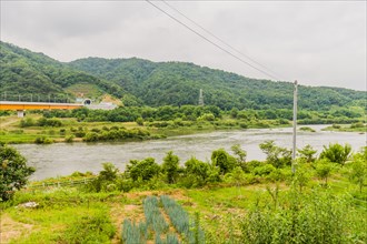 Overcast view of a river landscape with a train bridge in the distance and green mountains, in
