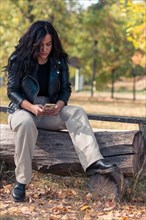 Serious Cheerful hispanic young Woman woman in leather jacket sitting on a bench in a park, looking