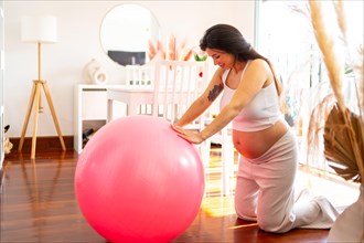 Domestic scene of a beauty pregnant woman using pink pilates ball to exercise at home