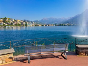 Bench on Lake Lugano and City with Water Fountain and Mountain in a Sunny Summer Day in Lugano,