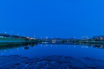 Blue hour scene of highway bridges reflected in calm water with city lights, in South Korea