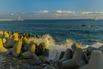 Waves crashing against tetrapods laying on ocean shoreline with two lighthouses on piers on horizon