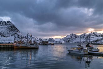Evening at a tranquil harbor with fishing boats and snow-covered mountains in the background,