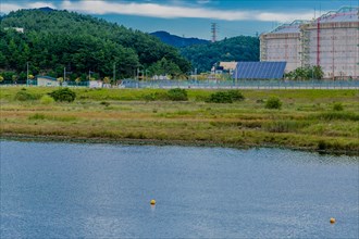 Industrial storage tanks beside a calm water body dotted with buoys, set against a mountain