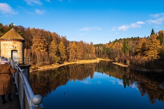 People walking along a dam, surrounded by colourful autumn trees under a blue sky, Ronsdorfer