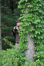 Huntress with rifle and binoculars behind a spruce (Picea) Allgaeu, Bavaria, Germany, Europe