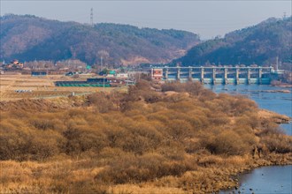 River cutting through a landscape with dam, houses, and hills under a clear sky, in South Korea