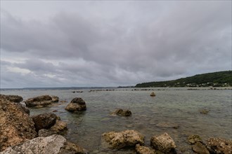 Seascape of rocky shoreline on a cloudy day with buildings on tree lined shore in the distance in