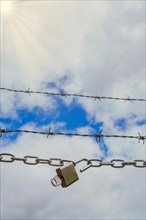 Chain with padlock open with key next to a barbed wire with a blue cloudy sky with sun rays,