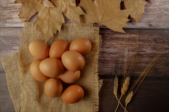Fresh eggs on a burlap cloth on a wooden table next to ears of wheat and dried leaves