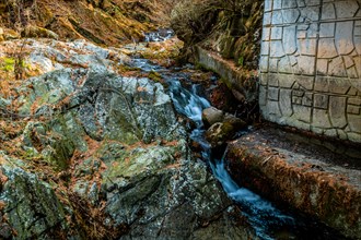 Water cascades over rocks in a stream next to a brick wall in a natural wooded landscape, in South