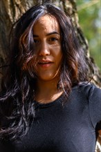 Smiling Cheerful hispanic young woman in black shirt standing in bright sunlight by a tree, blurred