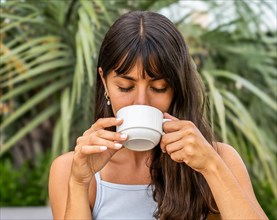 A woman is drinking coffee from a white cup with her eyes closed. She is wearing a tank top and has
