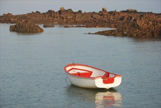 A lone red and white boat floating on calm sea water with rocks in the background