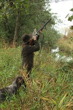 Female hunter aiming at flying mallard (Anas platyrhynchos) on the bank of a body of water during a