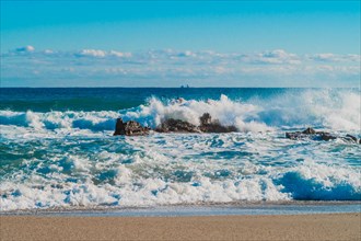 Crashing waves create spray around rocks on a sunlit beach with distant horizon, in South Korea