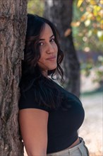 A relaxed Cheerful hispanic young latina sensual woman leaning against a tree trunk amidst nature,