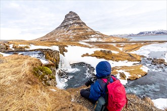 Adventurous photographer woman in winter in Iceland photographing sitting at Kirkjufell waterfall