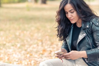 Latino Woman sitting on a park bench focused on her smartphone, wearing casual clothes, outdoors in