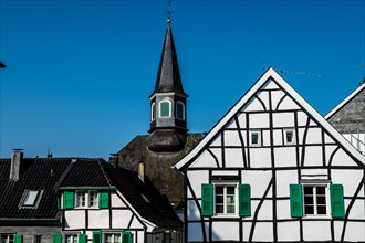 White half-timbered house with green shutters in front of a church tower under a blue sky,