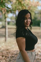 A smiling Cheerful hispanic young woman outdoors wearing a black shirt and beige pants with trees