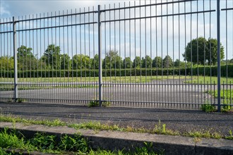 View through a high metal fence onto an empty car park surrounded by green space and trees,