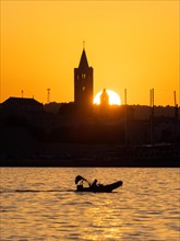 Motorboat in the golden evening light at sunset, silhouette of the church towers of Rab, town of