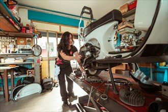 A woman mechanic works on a vintage scooter in a cluttered workshop, wearing black overall and