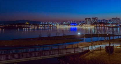 Night view of a lit pathway along a tranquil urban riverside with city lights, in South Korea