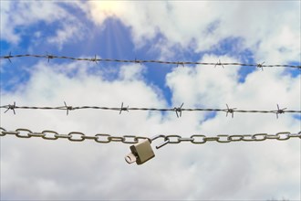 Chain with padlock open with key next to a barbed wire with a blue cloudy sky with sun rays,