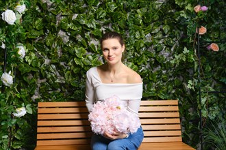 Cute woman sits on a wooden bench holding a bouquet of delicate pink flowers on her knees