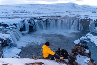 Adventurous woman in winter in Iceland sitting looking at frozen Godafoss waterfall