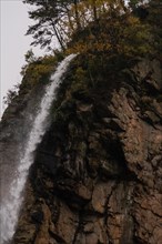 Vertical cliff face with a slender waterfall and scattered autumnal vegetation, in South Korea